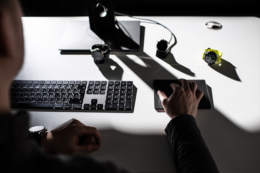 A person uses a touchpad at a white desk, surrounded by a keyboard and several Tag Heur Watches, with high contrast shadows.